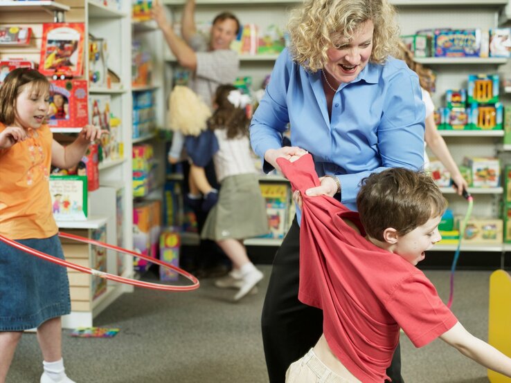 Ein Junge in rotem Shirt wird von einer blonden Frau in einem Spielzeugladen zurückgehalten, umgeben von spielenden Kindern und bunten Spielsachen.