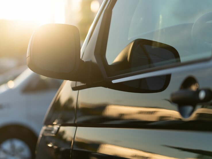 Explosionsgefahr bei bestimmten Gegenständen bei Hitze im Auto Ein parkendes schwarzes Auto, dessen Seitenspiegel von der untergehenden Sonne angestrahlt wird, daneben ein weißes Fahrzeug.