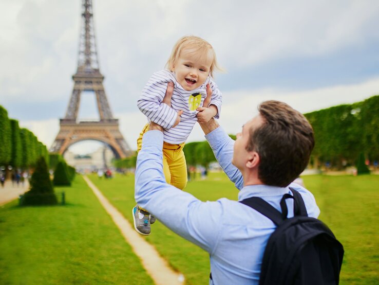 Vornamen, die von Städten inspiriert sind Ein lachender Vater hebt sein fröhliches Kleinkind vor dem Eiffelturm in Paris in die Luft, auf einer grünen Wiese.