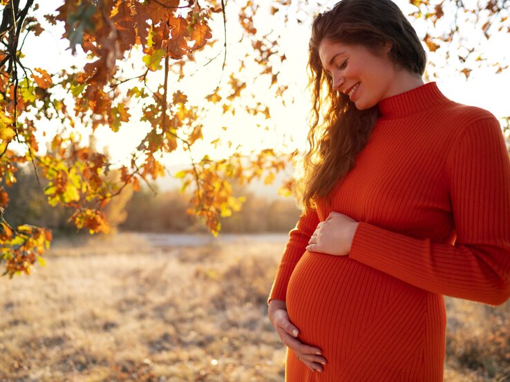 Vornamen Herbst, Namen Herbst, Mädchennamen Herbst, Jungennamen Herbst Junge schwangere Frau in einem orangefarbenen Kleid lächelt glücklich und hält ihren Bauch in einer herbstlichen Landschaft.
