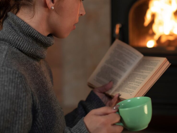 Junge Frau mit geflochtenem Zopf liest vertieft ein Buch vor einem Kaminfeuer und hält eine grüne Tasse in der Hand.