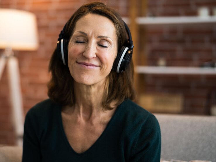 Eine lächelnde Frau mittleren Alters mit geschlossenen Augen genießt Musik über große Kopfhörer in einem gemütlichen Wohnzimmer.