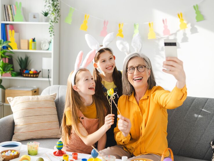 Ältere Frau mit grauen Haaren und Brille macht lachend ein Selfie mit zwei jungen Mädchen, alle tragen Hasenohren und Gelb.