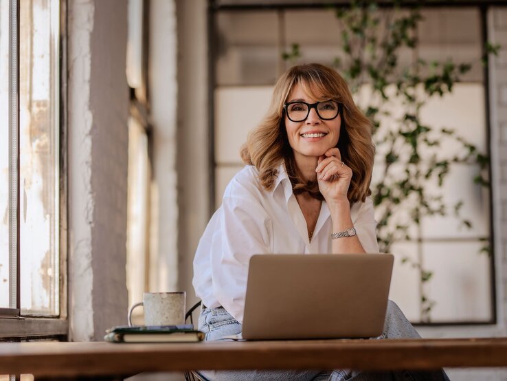 Eine lächelnde Frau mit schulterlangem Haar und Brille sitzt bei Hitze im hellen Büro entspannt am Laptop und blickt freundlich zur Kamera.