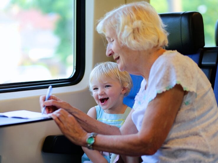 Lachendes blondes Kind sitzt neben einer fröhlichen Seniorin im Zug, die mit Stift und Papier auf dem Klapptisch schreibt.