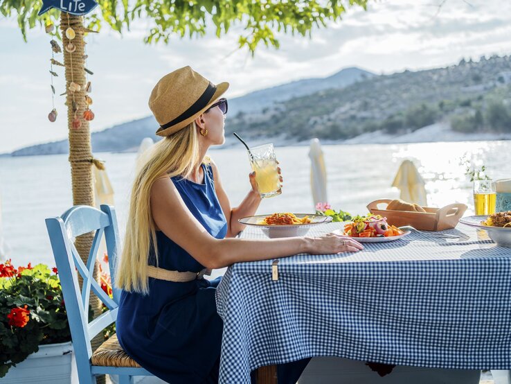 Eine blode Frau sitzt im Urlaub an einem reich gedeckten Tisch, nippt an einem Strohhalm und guckt in die Fern aufs Meer und angrenzende Berge. Es ist ein sonniger Nachmittag.