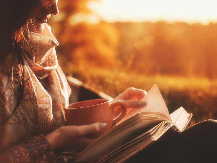 Eine Frau mit braunen Haaren liest bei goldenem Sonnenuntergang ein Buch und hält eine orangefarbene Tasse in der Hand.