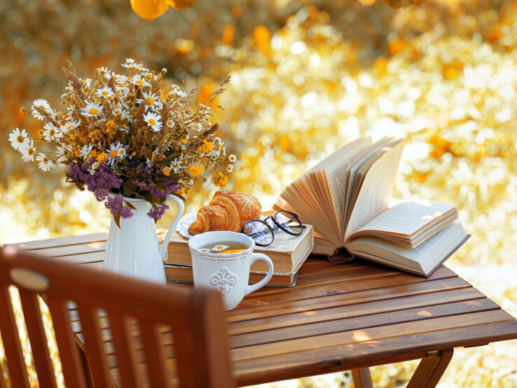 Ein Holztisch im warmen Herbstlicht mit Tee, Croissant, einem geöffneten Buch, einer Brille und einer Wildblumen-Vase.