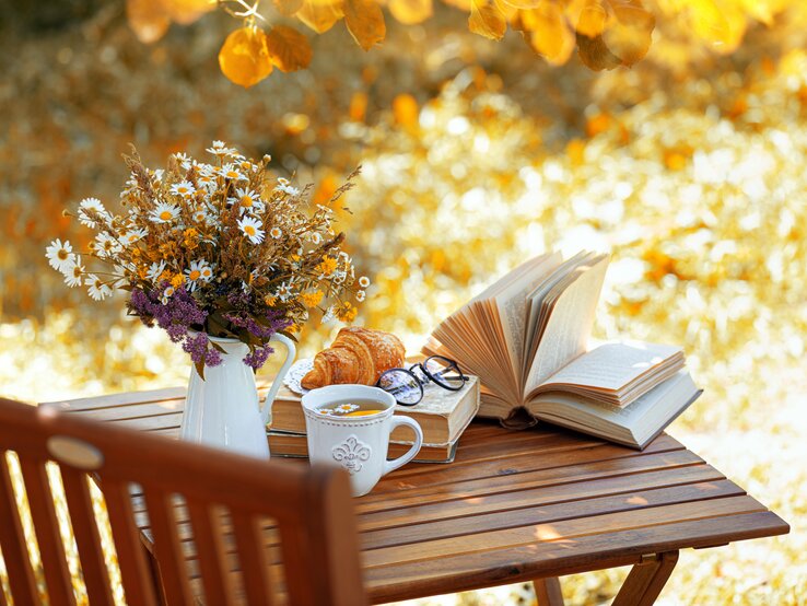 Ein Holztisch im warmen Herbstlicht mit Tee, Croissant, einem geöffneten Buch, einer Brille und einer Wildblumen-Vase.