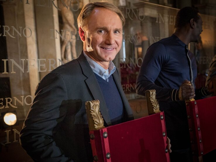 Autor Dan Brown lächelt in die Kamera, hinter einem roten Stuhl stehend.