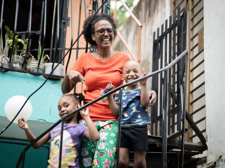 Junge Frau mit Brille umarmt zwei lachende Kinder auf einer Treppe vor bunt gestrichener Hauswand mit Pflanzen im Fenster.