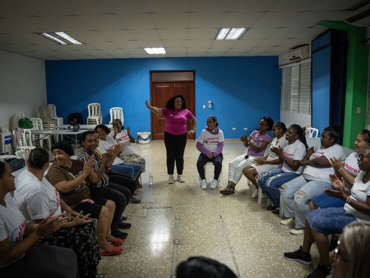 Frauen sitzen einander gegenüber in einem Stuhlkreis und applaudieren einer lächelnden Frau im pinken Shirt in der Raummitte. | © Martin Hanebeck