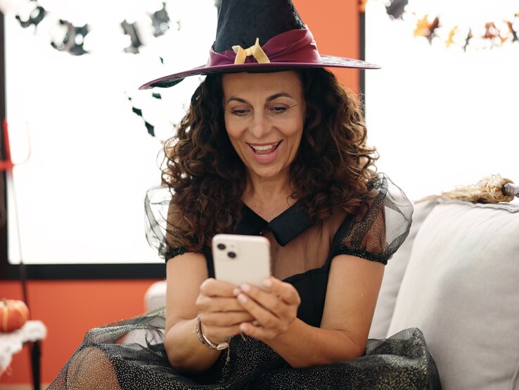 Halloween Sprüche Ein festlich dekorierter Raum mit Kürbissen, Kerzen und Spinnweben, dazu eine Person mit Hexenkostüm und Smartphone in der Hand.