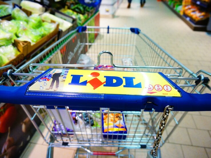 Einkaufswagen mit blauem Griff und Lidl-Logo im Supermarkt, im Hintergrund Gemüseabteilung und geflieste Böden sichtbar.