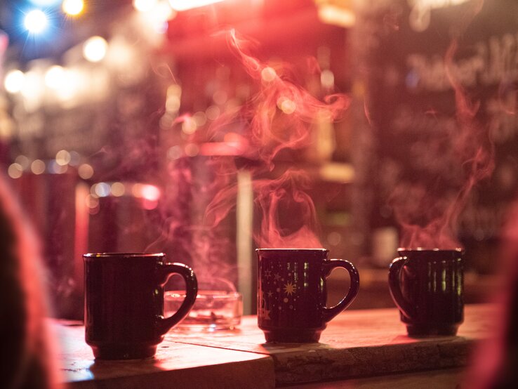 Drei dampfende Tassen Glühwein auf einem Holztisch, unscharfer Weihnachtsmarkt im Hintergrund mit Lichterglanz.