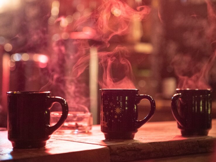 Drei dampfende Tassen Glühwein auf einem Holztisch, unscharfer Weihnachtsmarkt im Hintergrund mit Lichterglanz.