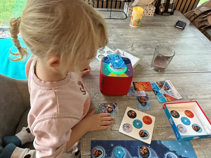 Kleines blondes Mädchen im rosa Pulli spielt konzentriert mit Disney-Karten neben roter Toniebox an einem Holztisch. | © Madeleine Liedke 