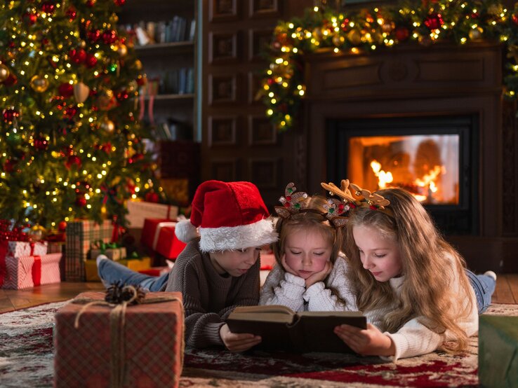 Drei Kinder lesen vertieft ein Buch vor dem Kamin, umgeben von Geschenken und festlich geschmücktem Weihnachtsbaum.