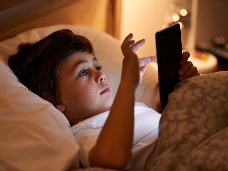Ein braunhaariger Junge im Grundschulalter liegt abends im Bett und blickt konzentriert auf ein Amazon Kindle Colorsoft Kids in seinen Händen.