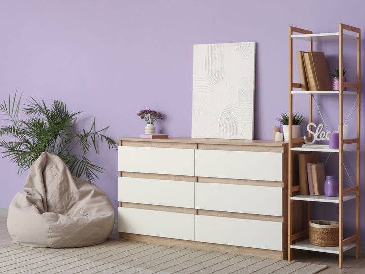 Modernes Wohnzimmer mit fliederfarbener Wand, Kommode aus Holz, Regal, Pflanzen, Sitzsack und ruhiger, heller Atmosphäre.