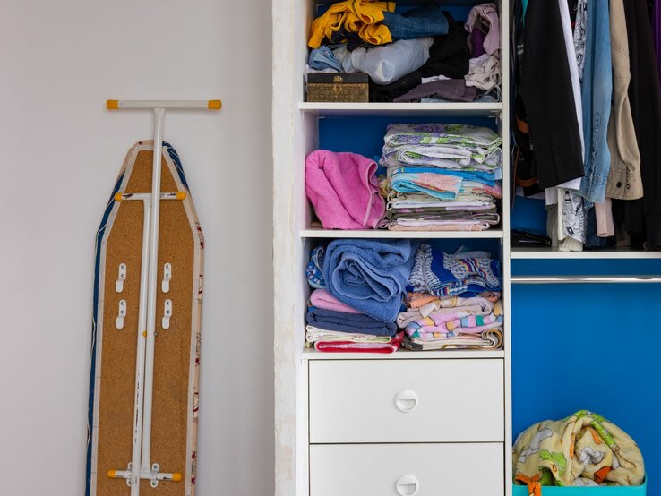 Ein geöffnetes Bügelbrett, das neben einem weißen Kleiderschrank mit einer blauen Tür steht. Im Schrank sind ordentlich gefaltete Handtücher und Bettwäsche auf Regalen sowie hängende Kleidungsstücke wie Jacken und Hosen zu sehen. Unter den Regalen befinden sich Schubladen, von denen zwei geschlossen sind und eine offen mit mehr Wäsche darin."