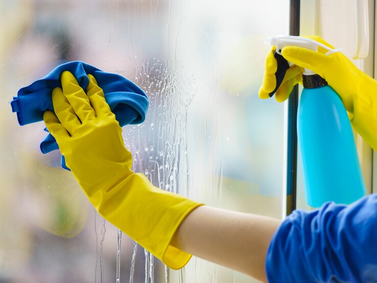 Behandschuhte Hände putzen Fenster mit Sprühflasche und Lappen