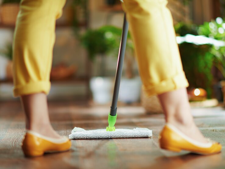  Das Bild zeigt die untere Hälfte einer Person beim Bodenwischen. Die Person trägt gelbe Hosen und passende gelbe Schuhe, was auf ein farblich abgestimmtes Outfit hinweist. Sie benutzt einen Swiffer Bodenwischer mit einem feuchten Bodentuch, das am Ende des Wischers angebracht ist, um den Holzboden zu reinigen. Die Szene spielt sich in einem hellen Raum mit Pflanzen im Hintergrund ab, was eine lebendige und gemütliche häusliche Umgebung andeutet. Der Fokus liegt auf der Wischaktion und dem Wischmop, was die einfache und effiziente Reinigung von Böden mit diesen modernen Reinigungswerkzeugen unterstreicht.