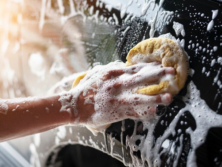 Das Foto zeigt einen Arm, der mit Seifenschaum bedeckt ist, während er mit einem gelben Schwamm ein Auto wäscht. Der Schaum bedeckt großflächig sowohl den Arm als auch das Auto, was auf eine gründliche Reinigung hindeutet. Das Bild ist in hellem Tageslicht aufgenommen, was die Frische und Sauberkeit der Szene unterstreicht.