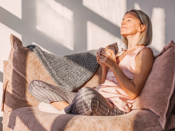 Dame mittleren Alters sitzt in Schlafanzug und mit Tasse in der Sonne.