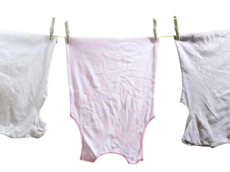 Drei vergilbte Wäschestücke, die an einer Wäscheleine hängen. In der Mitte befindet sich ein rosa Unterhemd, flankiert von zwei weißen Unterhemden, die jeweils mit grünen Wäscheklammern befestigt sind. | © iStock.com / MediaProduction
