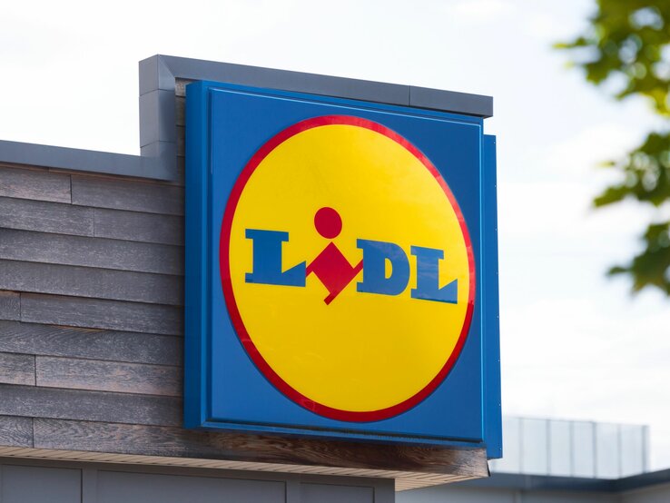 Das Bild zeigt das Firmenschild eines Lidl-Marktes. Im Mittelpunkt steht das markante Firmenlogo auf einem blauen Hintergrund, bestehend aus einem gelben Kreis mit einem roten Rand, in dem der Name "Lidl" in blauen Buchstaben steht. Innerhalb des gelben Kreises befindet sich das stilisierte Bild einer Person, die einen Einkaufswagen schiebt. Das Schild ist an der Seite eines Gebäudes mit einer hölzernen Fassade befestigt. Im Hintergrund sieht man einen leicht bewölkten Himmel und das Laub eines Baumes in der Ecke oben rechts. Das Bild wirkt klar und modern, typisch für die Außengestaltung eines Supermarktes der Kette.