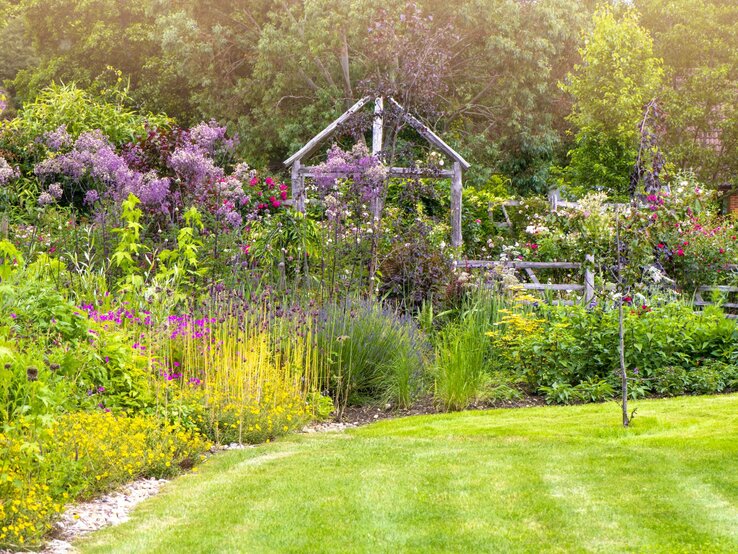  Das Bild zeigt ein üppiges Staudenbeet in einem Garten. Der Vordergrund ist von einem sorgfältig gemähten, grünen Rasen geprägt. Im Beet selbst ist eine Vielfalt von Pflanzen und Blumen in verschiedenen Höhen und Farben zu erkennen, darunter lila, rosa, gelbe und rote Blüten. Eine Holzpergola, die mit kletternden Pflanzen bewachsen ist, bildet einen natürlichen Rahmen im Hintergrund. Die Komposition der Pflanzen und die Gestaltung des Gartens lassen auf eine durchdachte Gartengestaltung schließen, möglicherweise inspiriert von einem Cottage-Garten, mit einem Fokus auf Vielfalt und Natürlichkeit.