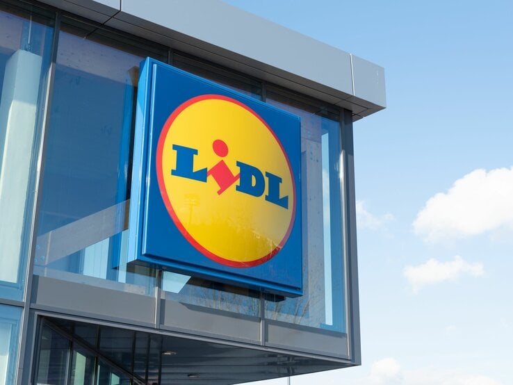  Das Bild zeigt das Logo des Einzelhandelsunternehmens Lidl an der Außenfassade eines Gebäudes. Das Logo ist in den charakteristischen Unternehmensfarben Blau und Gelb gehalten, mit einem roten Akzent, der den Teil des „i“ im Wort „Lidl“ darstellt. Der Hintergrund ist ein klarer Himmel mit einigen Wolken, was auf einen Standort des Geschäfts bei Tageslicht hinweist.