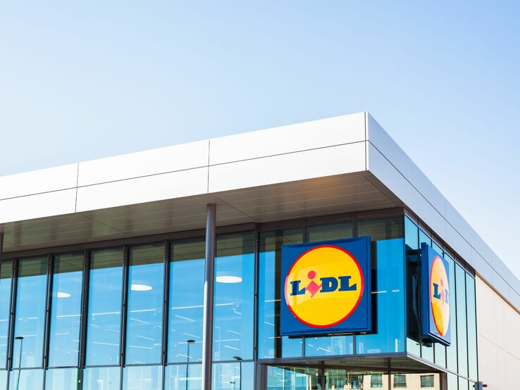  Das ist ein Bild eines modernen Gebäudes mit einer großen Glasfassade. Auf der Fassade sind zwei große, quadratische Schilder mit dem Logo von Lidl, einer bekannten Supermarktkette, zu sehen. Das Logo besteht aus einem blauen Hintergrund mit einem gelben Kreis, in dem sich der Name "Lidl" in roten Buchstaben befindet. Darüber ist ein kleiner, stilisierter Kopf mit einem gelben Umriss abgebildet. Das Gebäude selbst hat eine klare, rechtwinklige Form mit einem flachen Dach, was typisch für kommerzielle Gebäude dieser Art ist.