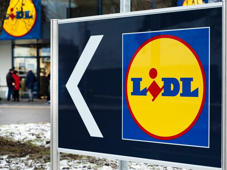  Das Bild zeigt ein großes Schild mit einem Pfeil und dem Logo des Supermarkts Lidl. Der Pfeil weist nach links, was darauf hinweist, dass die Filiale in diese Richtung liegt. Im Hintergrund sieht man den Eingangsbereich eines Lidl-Marktes mit einigen Personen, die sich in der Nähe befinden. Der Boden ist teilweise mit Schnee bedeckt, was auf winterliche Wetterbedingungen schließen lässt.