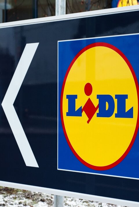  Das Bild zeigt ein großes Schild mit einem Pfeil und dem Logo des Supermarkts Lidl. Der Pfeil weist nach links, was darauf hinweist, dass die Filiale in diese Richtung liegt. Im Hintergrund sieht man den Eingangsbereich eines Lidl-Marktes mit einigen Personen, die sich in der Nähe befinden. Der Boden ist teilweise mit Schnee bedeckt, was auf winterliche Wetterbedingungen schließen lässt.