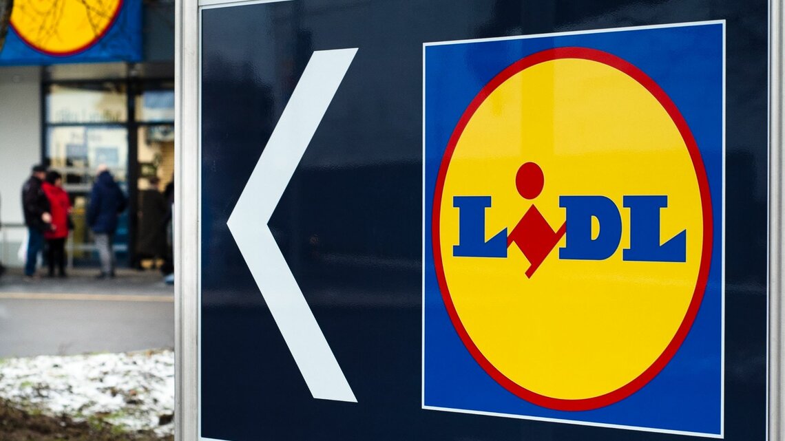  Das Bild zeigt ein großes Schild mit einem Pfeil und dem Logo des Supermarkts Lidl. Der Pfeil weist nach links, was darauf hinweist, dass die Filiale in diese Richtung liegt. Im Hintergrund sieht man den Eingangsbereich eines Lidl-Marktes mit einigen Personen, die sich in der Nähe befinden. Der Boden ist teilweise mit Schnee bedeckt, was auf winterliche Wetterbedingungen schließen lässt.