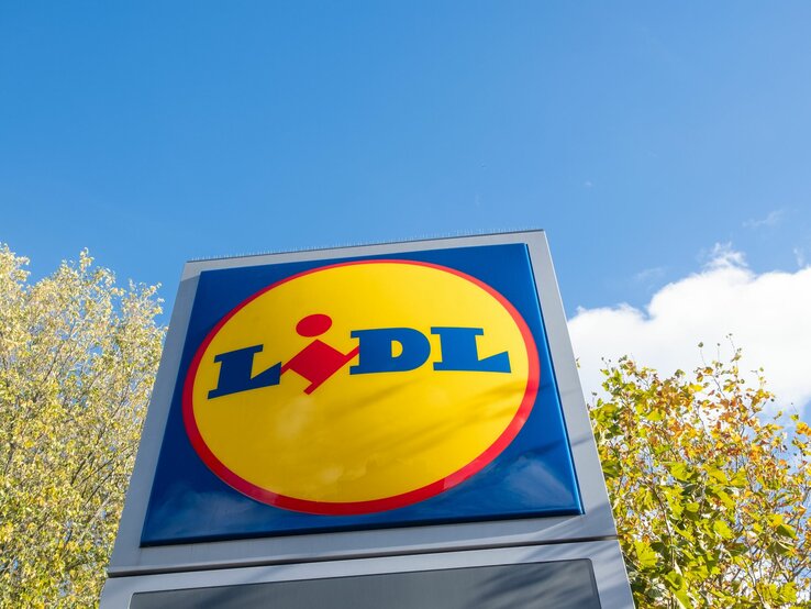  Das Bild zeigt das Logo von Lidl auf einem großen Schild, das gegen den Himmel und einige Baumkronen im Hintergrund steht. Das Logo besteht aus einem blauen Rechteck mit abgerundeten Ecken, in dem der Name "Lidl" in weißer Schrift steht.