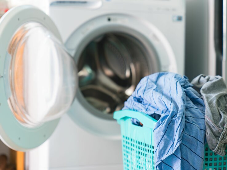 Geöffnete Waschmaschine mit frischer Wäsche neben voller blauer Wäschekorb.