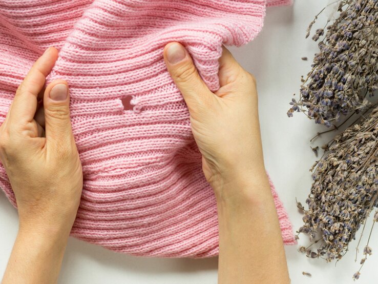 Hände halten rosa gestrickten Pullover neben getrocknetem Lavendel auf weißem Untergrund.