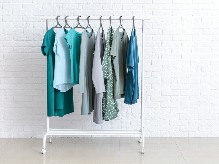 Kleiderstange mit blauen und grünen Kleidungsstücken vor weißer Ziegelwand.