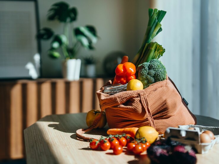 Stoffbeutel mit frischem Gemüse und Obst auf Holztisch, Sonnenlicht, Zimmerpflanzen im Hintergrund.