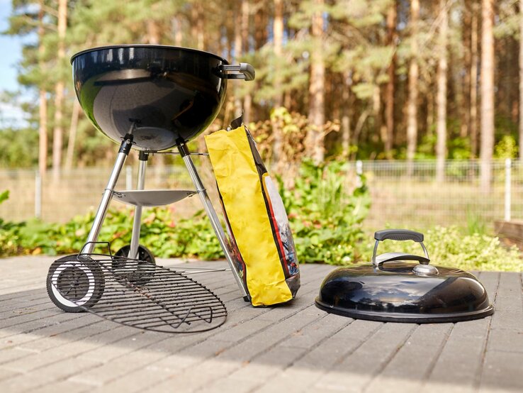 Schwarzer Kugelgrill bereit zum Anfeuern auf Terrasse mit Tüte Holzkohle, im Hintergrund Kiefernwald.