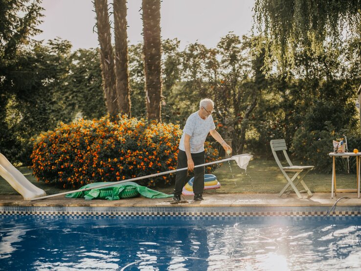 Senior mit grauem Haar entfernt Blätter aus einem Pool, umgeben von leuchtenden Blumen.