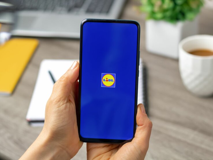 Eine Hand hält ein Smartphone mit Lidl-Logo auf blauem Bildschirm, im Hintergrund unscharfe Büroelemente.