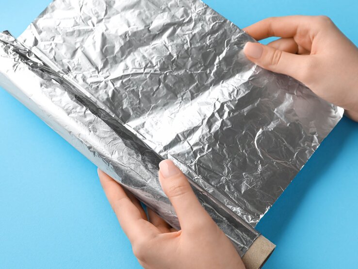 Zwei Hände, die eine Rolle Aluminiumfolie abrollen. Die Aluminiumfolie ist silbern und leicht zerknittert, und sie wird gegen einen blauen Hintergrund gehalten.
