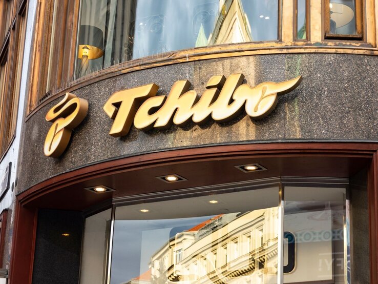 Tchibo Haus und Garten Artikel Eingang einer Tchibo-Filiale mit großem goldenen Logo auf dunkler Fassade und spiegelnder Glasscheibe.