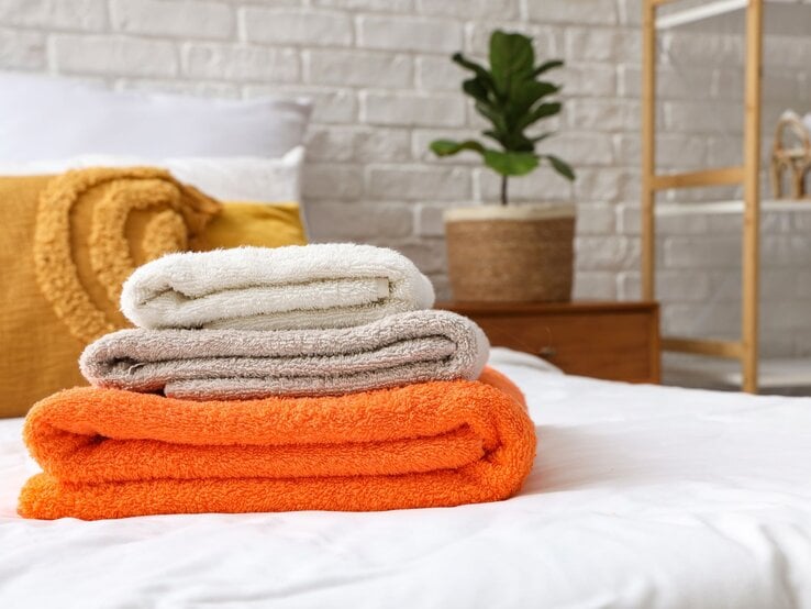 Drei gefaltete Handtücher in Weiß, Beige und Orange liegen gestapelt auf einem Bett, im Hintergrund eine Pflanze vor weißer Backsteinwand.