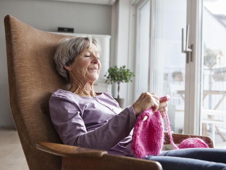 richtig Heizen für mollige Wärme Eine ältere Frau sitzt lächelnd in einem Sessel und strickt. Sie genießt die Wärme in der Wohnung.