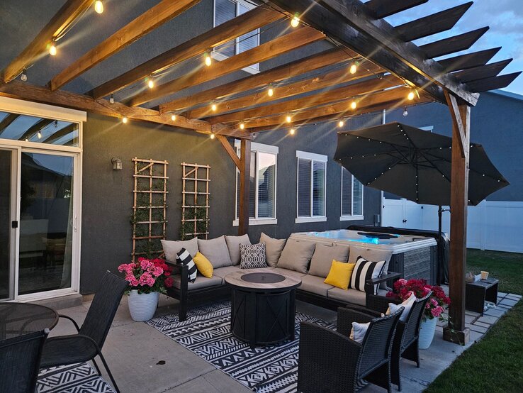 Gemütliche überdachte Terrasse am Abend mit Lichterketten, Lounge-Sofa, dekorativen Kissen, Feuertisch und beleuchtetem Whirlpool im Hintergrund.